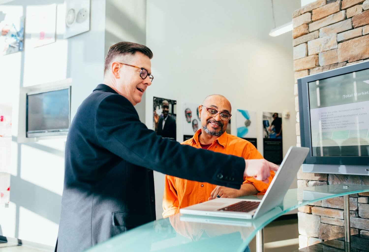 Two men in an office setting looking at a laptop, with one pointing to the screen and the other smiling.